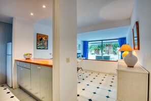A kitchen with white cabinets, a wooden countertop, and a bowl of fruit. There is a window with blue curtains and a view of greenery outside.