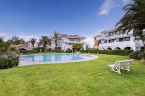 A swimming pool surrounded by well-maintained grass and palm trees, with a white bench and a building with balconies in the background. - Magnificent apartment with pool 2 minutes from Pals beach (Pals)
