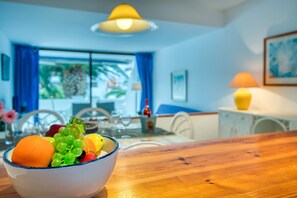 A dining area with a wooden table, a bowl of fruit, and a view of the outdoors through a window with blue curtains.
