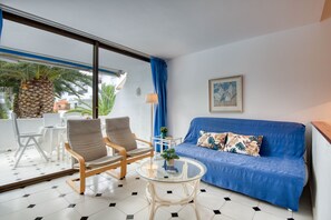 A room with a blue sofa, two wooden chairs, a glass table, and a view of the outdoors.