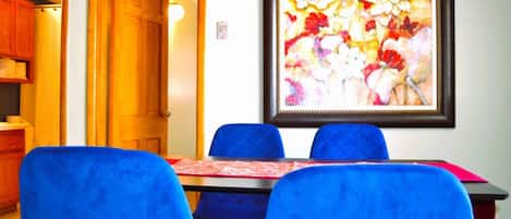 A dining area with a table and blue chairs, a framed floral artwork on the wall, and a wooden door leading to another room.