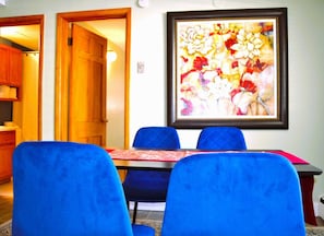 A dining area with a table and blue chairs, a framed floral artwork on the wall, and a wooden door leading to another room.