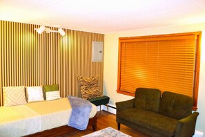 Una habitación con una cama, un sillón verde, un muro de listones de madera y una ventana con persianas.