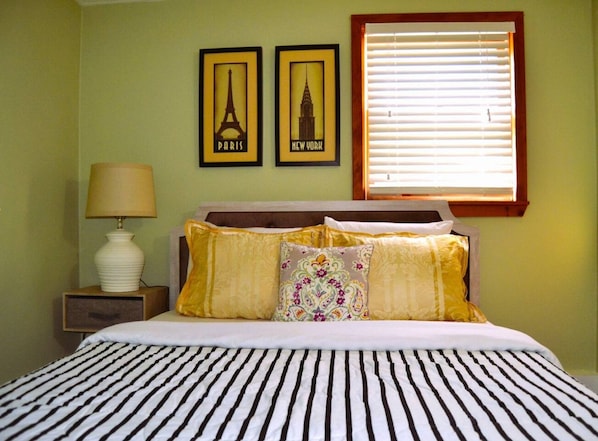 A bed with striped bedding, a nightstand with a lamp, and two framed pictures of Paris and New York on the wall.