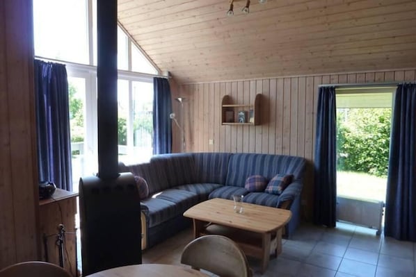 A cozy cabin interior with a wooden ceiling, a large window, a sofa, a table, and a built-in wood stove.
