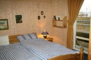 Ett litet, ombonat rum med en säng i trä, ett nattduksbord med en lampa och ett fönster med gardiner.