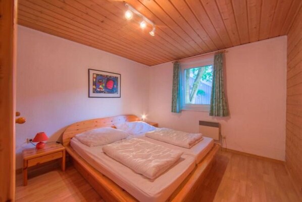 Une chambre avec un plafond en bois, un lit avec deux oreillers, une table de chevet avec une lampe et une fenêtre avec des rideaux.