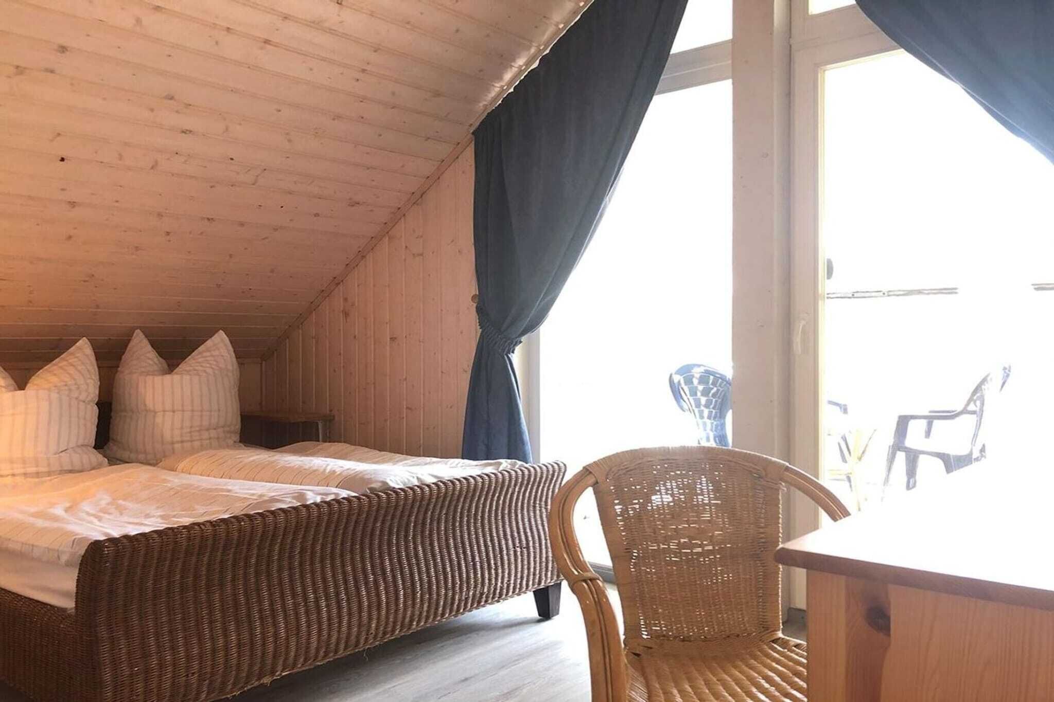 Una habitación con techo inclinado, dos camas con almohadas, una silla de mimbre y una mesa de madera.