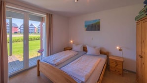 A bedroom with two beds, a sliding glass door to a garden, a painting on the wall, and a wooden wardrobe.
