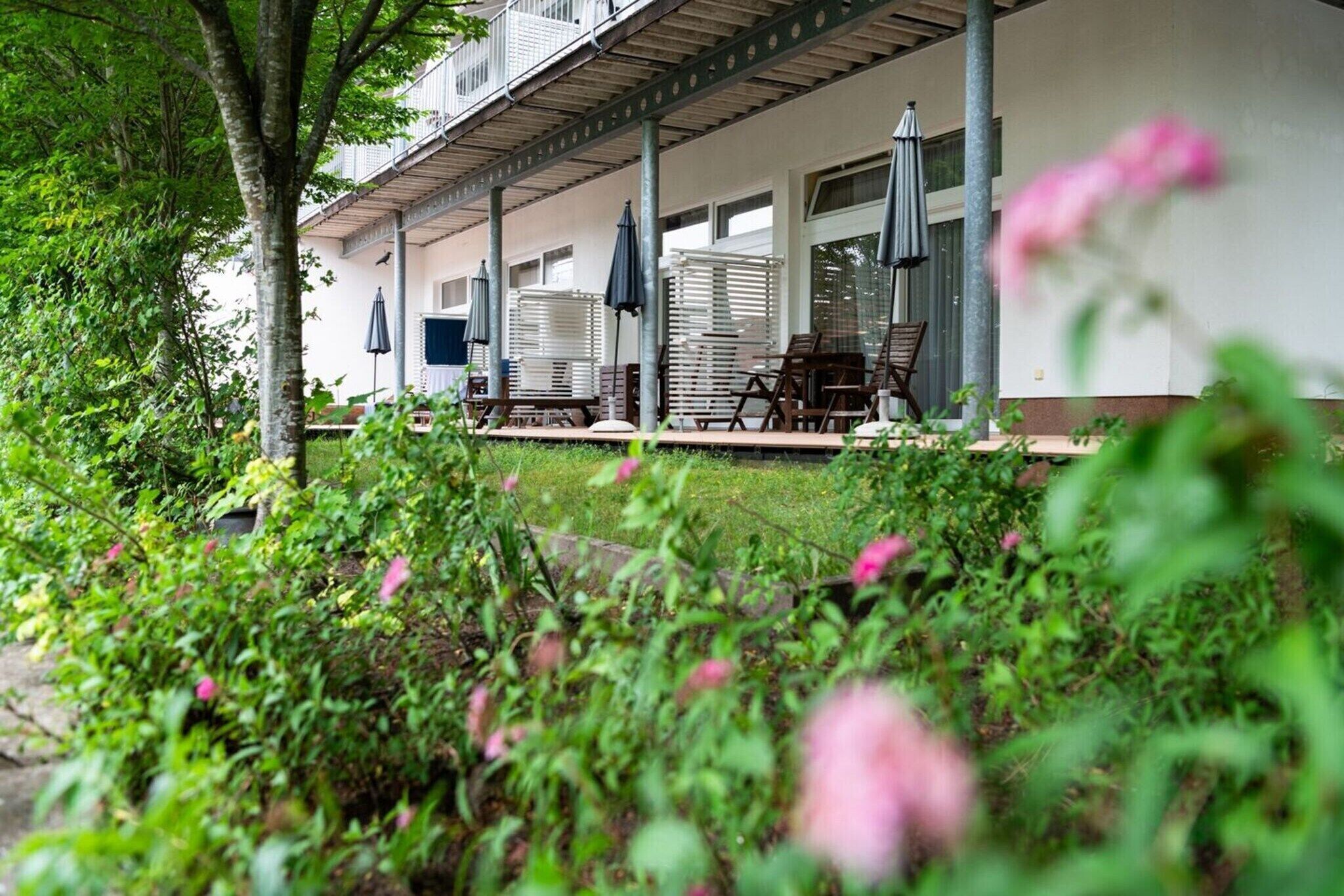 A covered outdoor seating area with chairs and umbrellas, surrounded by lush greenery and flowering plants.
