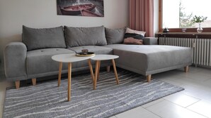 A modern living room with a grey sectional sofa, two small white side tables, a patterned rug, and a large window with curtains.