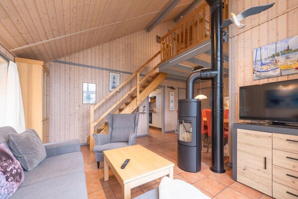A cozy living room with a sofa, a table, a TV, a fireplace, and a staircase leading to an upper level. - Ferienhaus im skandinavischen Stil (Rechlin)