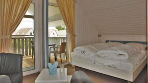A bedroom with a bed, a chair, a small table, and a balcony with a view of houses.