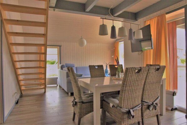 A dining area with a white table and wicker chairs, a wooden ladder, and a TV mounted on the wall.