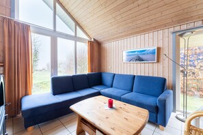 A modern living room with a blue sectional sofa, a wooden coffee table, and a large window with curtains. - Ferienhaus im Ferienpark Mueritz (Rechlin)