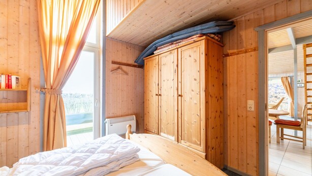 Una habitación de madera con una cama, un armario y vistas a través de un ventanal.