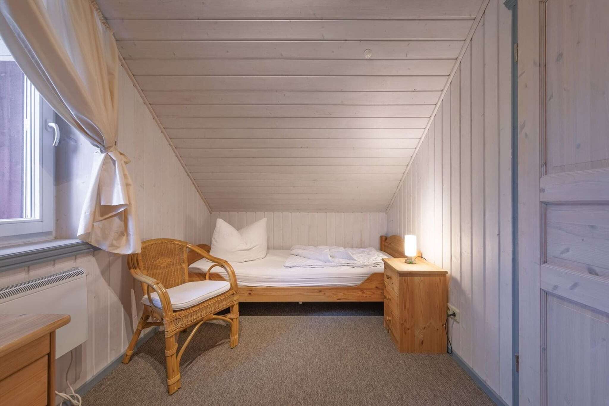 Una habitación pequeña con cama individual, techo inclinado, una silla de madera, una mesita de noche con lámpara y un radiador.