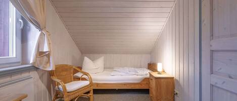 Una habitación pequeña con cama individual, techo inclinado, una silla de madera, una mesita de noche con lámpara y un radiador.