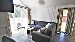 Ein modernes Wohnzimmer mit grauem Sofa, einem kleinen Tisch und einem Sessel. An der Wand ist ein Fernseher angebracht, und der Raum hat Holzdielen sowie große Fenster mit Vorhängen.