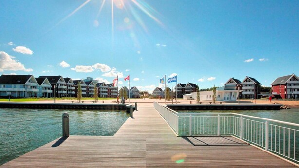 Ein Hotel am Wasser mit einer hölzernen Anlegestelle, Flaggen und modernen Gebäuden, strahlend blauer Himmel.