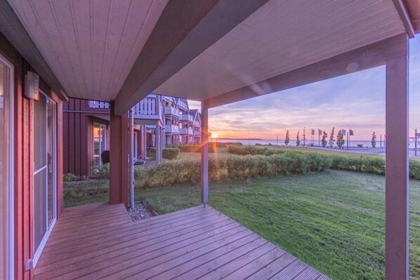 A wooden deck with a view of a sunset over a grassy area and a row of buildings in the background.