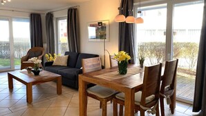A modern living room with a sofa, dining table, and chairs. There is a large window with curtains, a framed picture on the wall, and a vase with flowers on the table. - liebevoll eingerichtetes Appartement (Rechlin)