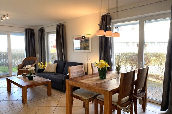 Un salon moderne avec un canapé, une table à manger et des chaises. Il y a une grande fenêtre avec des rideaux, un tableau encadré au mur et un vase rempli de fleurs sur la table.