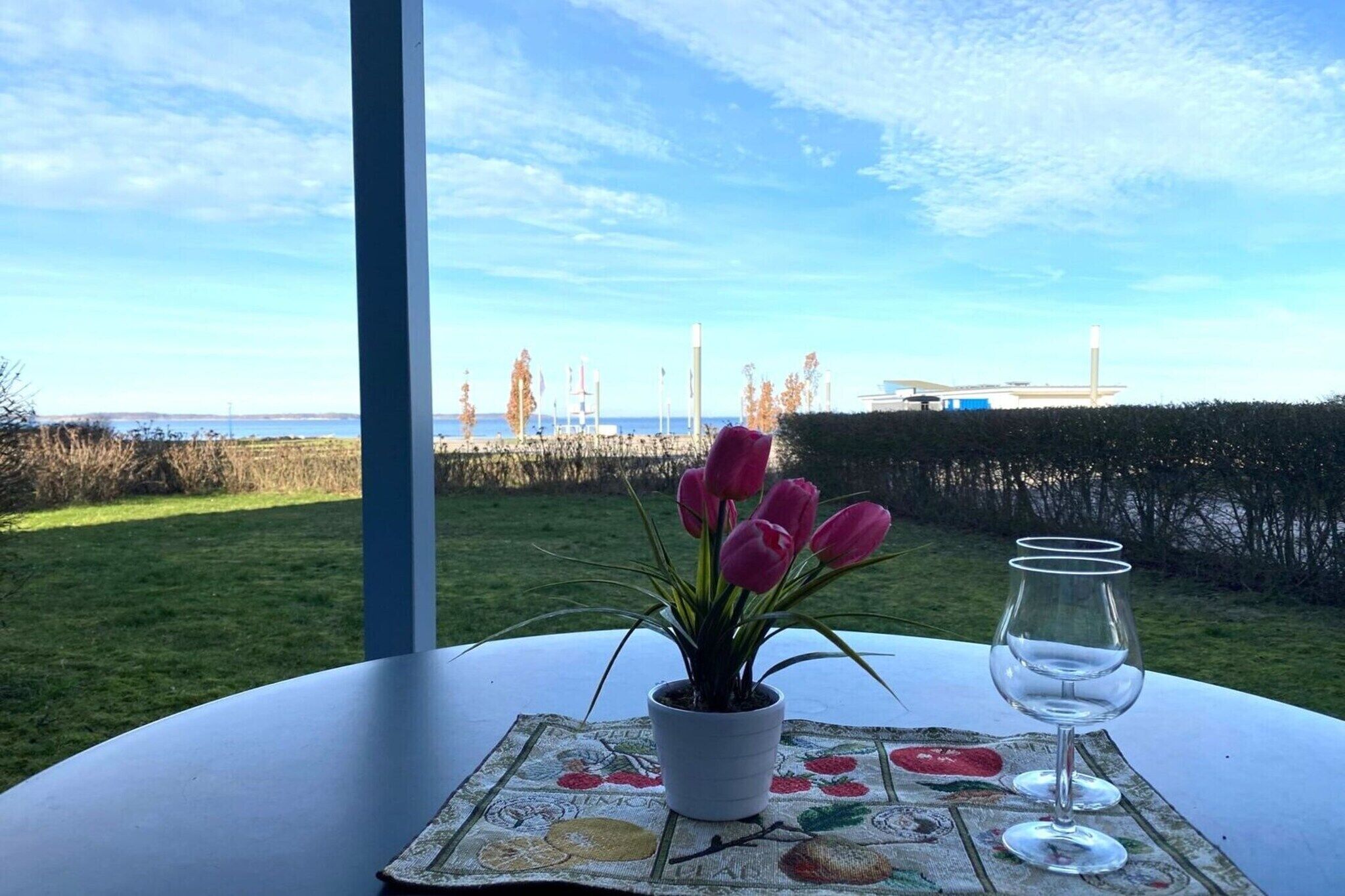 A table with a potted plant and a wine glass, set on a tablecloth with fruit designs, overlooking a grassy area and a body of water.