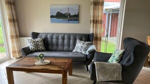 Een woonkamer met een donkergrijze bank, een houten bijzettafeltje en een stoel met een plaid. Een schilderij van een vuurtoren hangt aan de muur.
