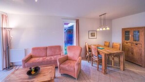 Un salon comprenant un canapé, un fauteuil, une table à manger en bois et des chaises. On y trouve également un radiateur, un lampadaire et un tableau accroché au mur.