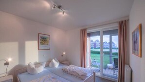 A hotel room with two beds, a large window with curtains, and a view of a building and greenery outside. - liebevoll eingerichtetes Appartement (Rechlin)