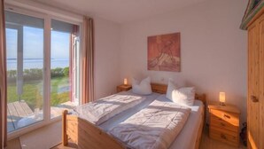 A bedroom with a large window, a bed, a nightstand, and a wooden wardrobe.