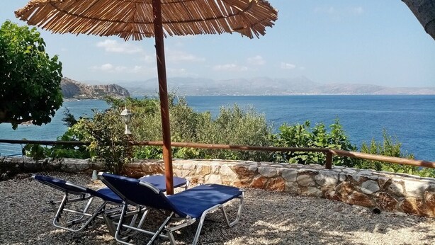 A beachside setting with two sun loungers under a thatched umbrella, a stone wall, and a view of the sea and distant mountains. - 2 Annexed apartments over the sea – Private place to swim (ravdoucha)
