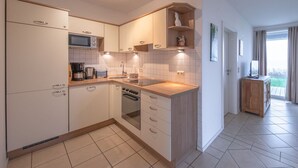 Una cocina moderna con armarios blancos, microondas, estufa y lavabo. Hay una encimera de madera y un mueble pequeño de madera. La cocina tiene suelo de baldosas y una ventana con cortinas.