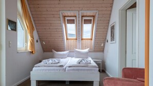 Een slaapkamer met een houten plafond, twee ramen, een bed met witte lakens, een nachtkastje en een roze stoel.