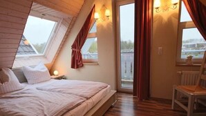 A bedroom with a sloped ceiling, a bed, a chair, and two windows with curtains.