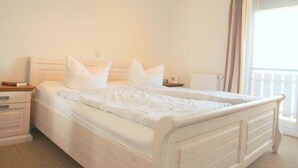 A neatly made bed with white linens and pillows, a bedside table with a lamp, and a window with curtains. - liebevoll eingerichtetes Apartement (Rechlin)