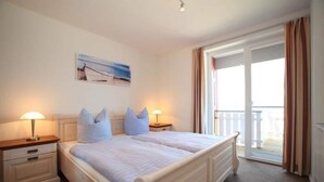 Un llit doble amb capçalera, dues taules de nit amb làmpades, un quadre a la paret i un balcó amb cortines.