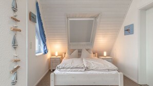 Une chambre avec un plafond en pente, un lit recouvert de linge de lit blanc, deux tables de chevet avec des lampes, une fenêtre avec des rideaux bleus et des décorations murales.