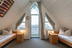 A cozy attic bedroom with two single beds, a wooden dresser, a clock, and a view of a white fence through a large window. - exklusives Appartement in Rechlin (Rechlin)