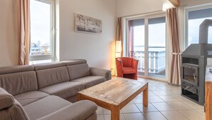A living room with a sofa, a wooden coffee table, a red chair, a fireplace, and large windows with curtains. - Appartement im Ferienpark in Rechlin (Rechlin)