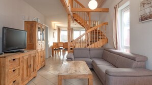 A spacious living room with a wooden staircase, a television on a wooden cabinet, and a large gray sofa. - Appartement im Ferienpark in Rechlin (Rechlin)