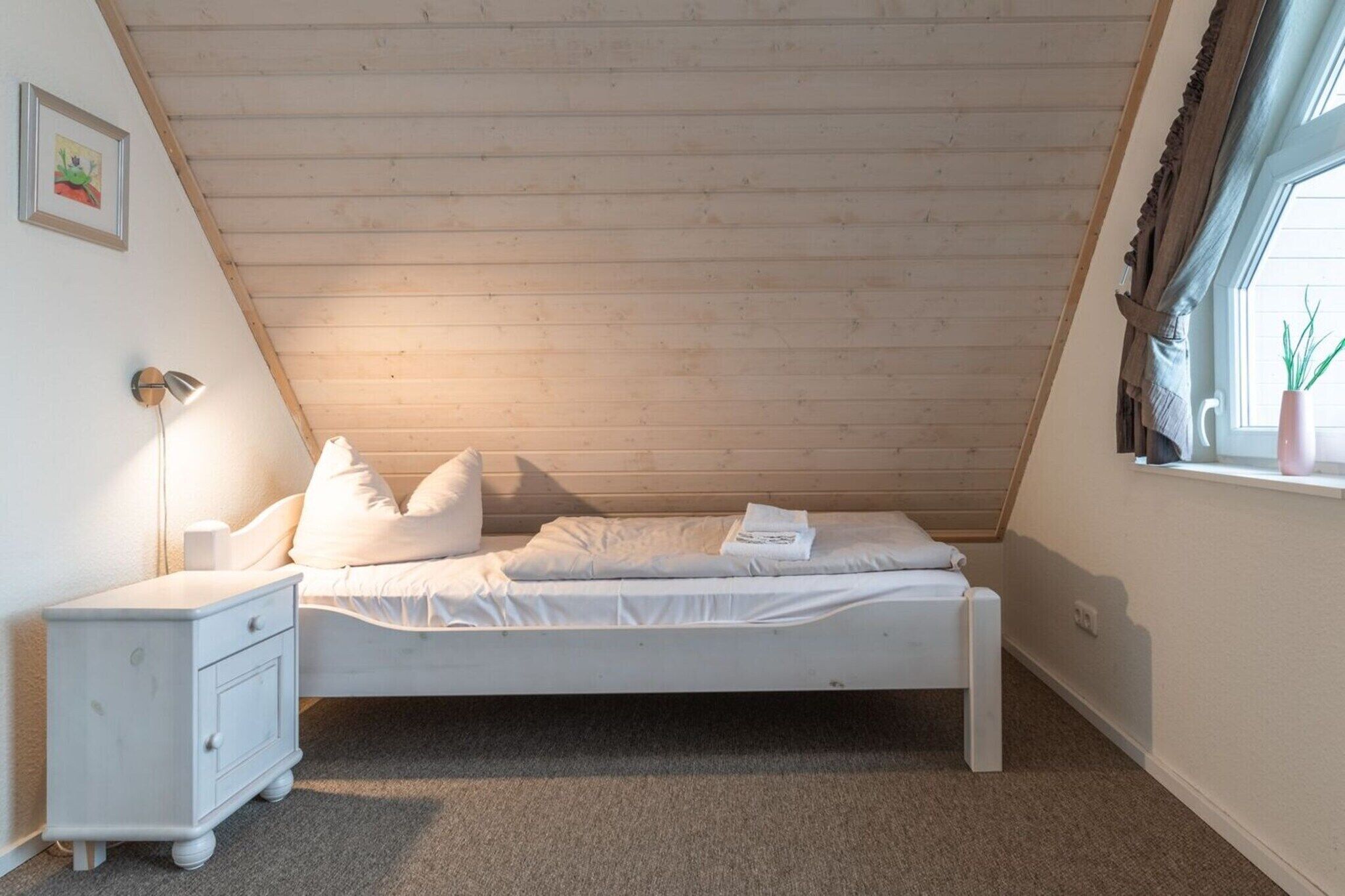 Quarto pequeno com cama de solteiro, teto inclinado, luminária de cabeceira, mesa de cabeceira e janela com cortinas.