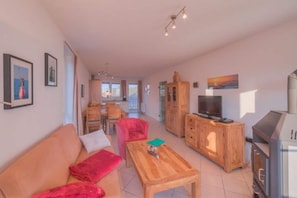 Un vaste espace de vie comprenant un canapé, un fauteuil rouge, une table basse en bois et un téléviseur posé sur un meuble en bois.
