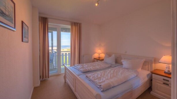 A bedroom with a double bed, two bedside tables with lamps, a sliding glass door leading to a balcony, and a framed picture on the wall. - Appartement im Ferienpark in Rechlin (Rechlin)