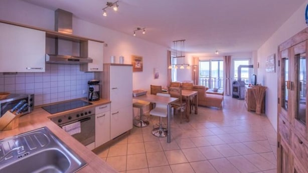 A modern kitchen with a gas stove, a coffee machine, and a sink. There is a dining area with a table and chairs. - Appartement im Ferienpark in Rechlin (Rechlin)
