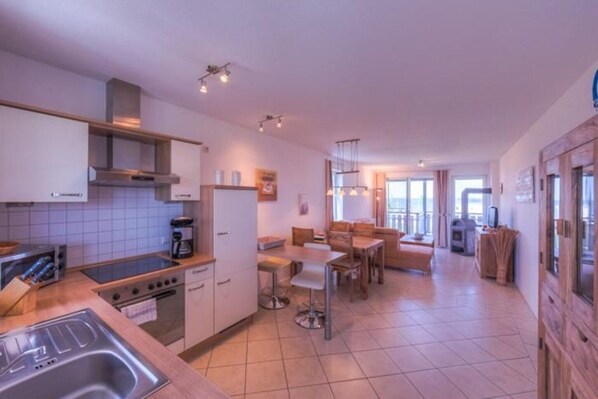 A modern kitchen with a gas stove, a coffee machine, and a sink. There is a dining area with a table and chairs. - Appartement im Ferienpark in Rechlin (Rechlin)