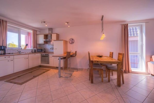 A modern kitchen with white cabinets, a dining table, and chairs. There is a window with curtains, a clock on the wall, and a hanging light fixture. - exklusives Appartement in Rechlin (Rechlin)