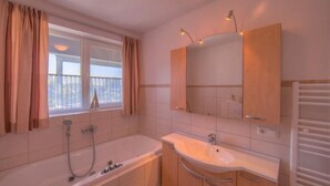 A bathroom with a bathtub, a sink, and a mirror. There is a window with curtains and a view of buildings outside. - Appartement im Ferienpark in Rechlin (Rechlin)