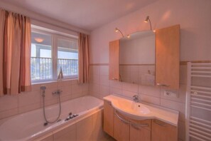 A bathroom with a bathtub, a sink, and a mirror. There is a window with curtains and a view of buildings outside. - Appartement im Ferienpark in Rechlin (Rechlin)